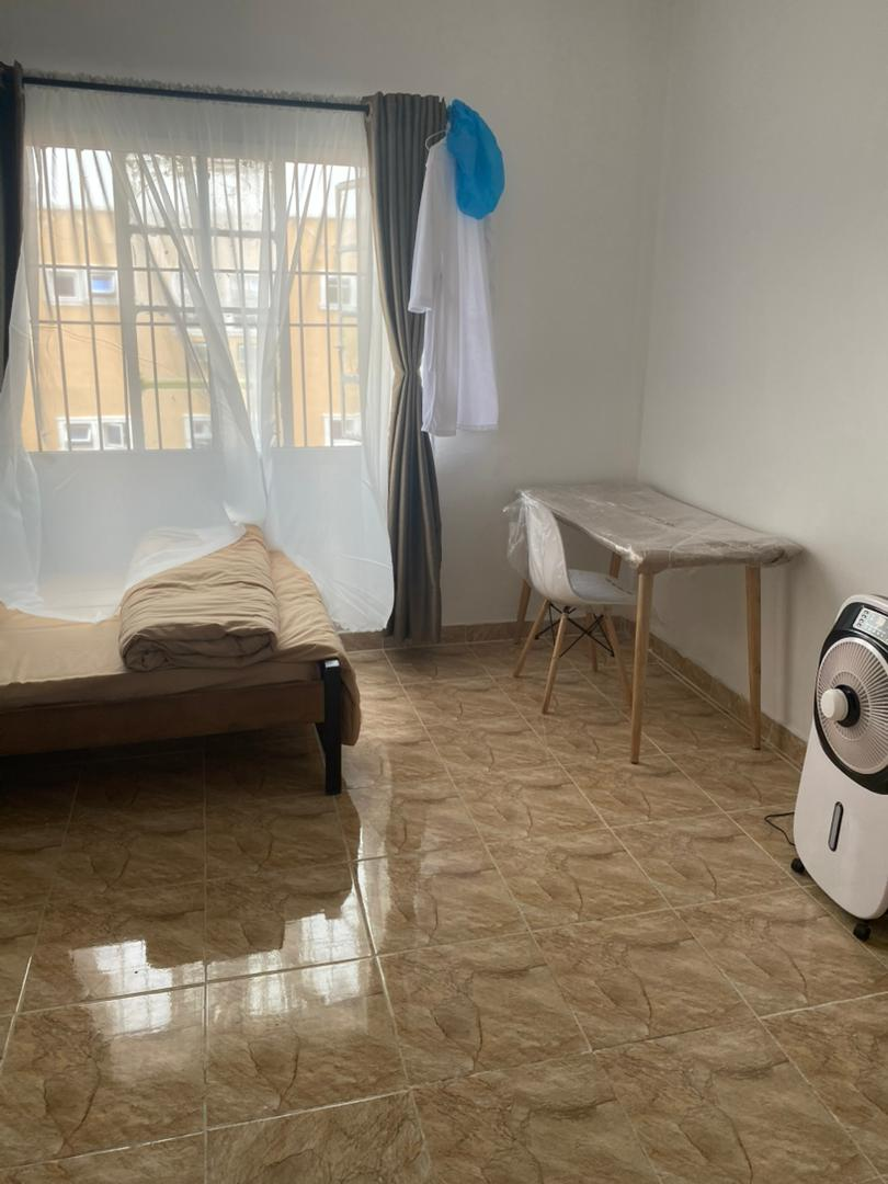 Bright single room with air cooler fan, desk, chair and single bed at Rilwan AJALA Homes